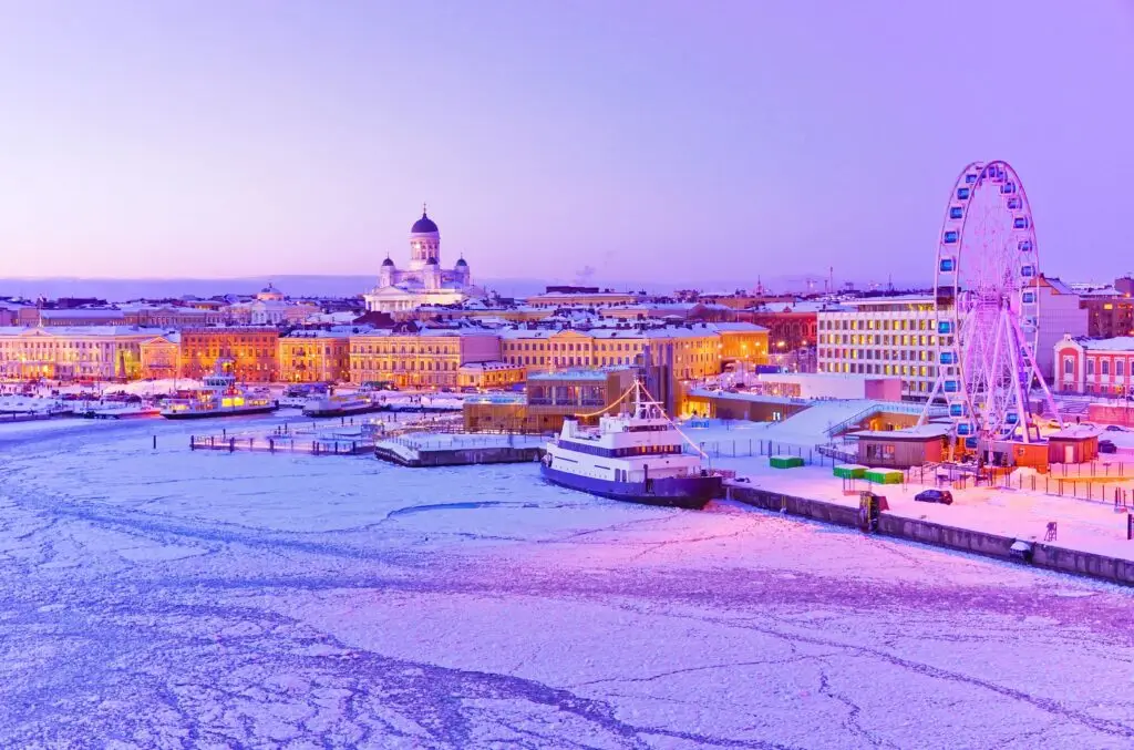 Icy winter view of Helsinki harbor at dusk with ferris wheel, frozen sea, and Helsinki Cathedral illuminated in the background