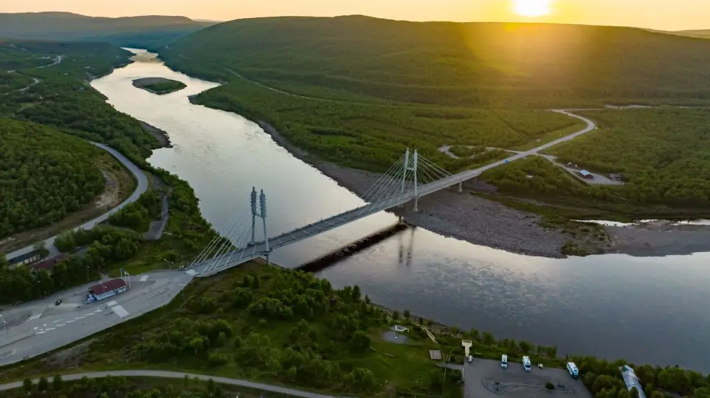 Aerial view of the Sami Bridge crossing the Teno River between Finland and Norway during summer, surrounded by lush green landscape and evening sunlight, promoting Northern Europe private tours and scenic travel experiences