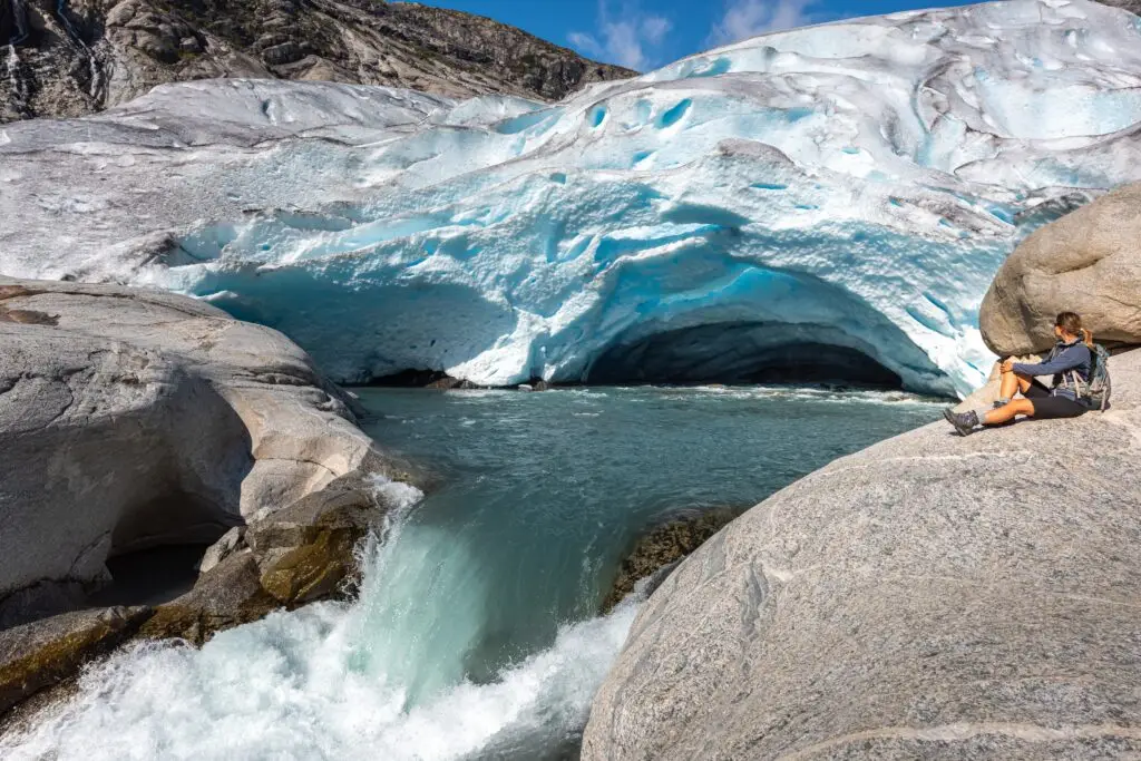 Hiker sitting on rocks beside the blue ice cave of Nigardsbreen Glacier in Norway, with glacial river and scenic mountain landscape in summer