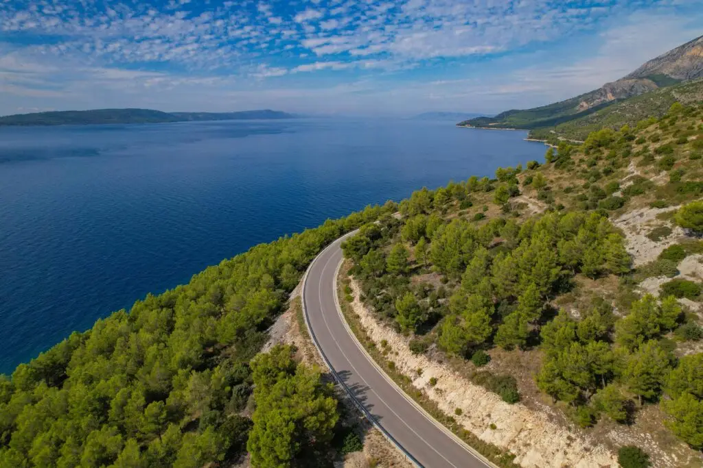 Aerial panoramic view of a scenic coastal highway along the Adriatic Sea in Croatia, surrounded by pine forests and turquoise waters, perfect for European road trips and private tours.