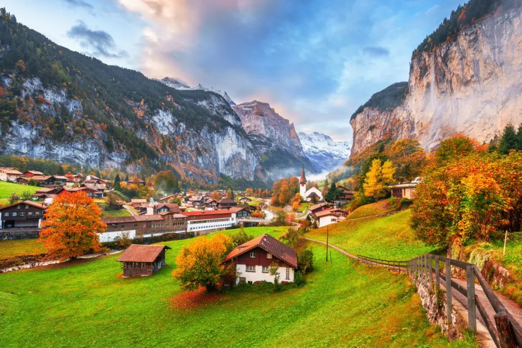 Scenic view of Lauterbrunnen Valley in Switzerland with alpine village houses, church steeple, green meadows, autumn foliage, and dramatic mountain cliffs in the Swiss Alps
