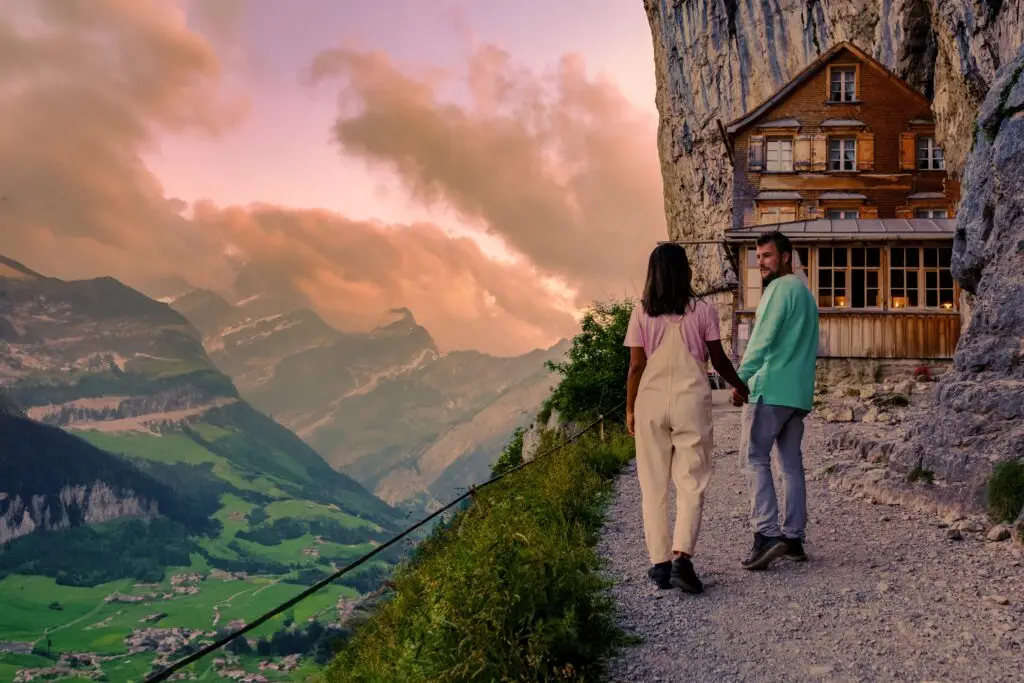 Couple walking toward Berggasthaus Aescher mountain guesthouse in the Swiss Alps at sunset, Appenzell, Switzerland