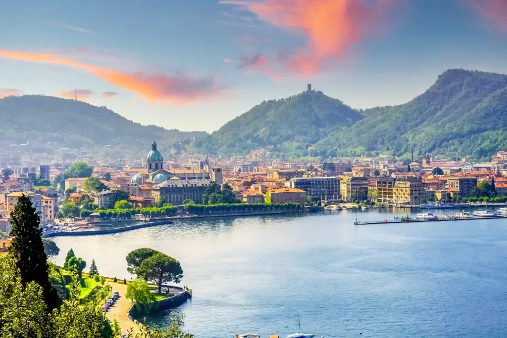 Scenic view of Lake Como and the historic city of Como, Italy, with mountains, waterfront buildings, and colorful sunset sky