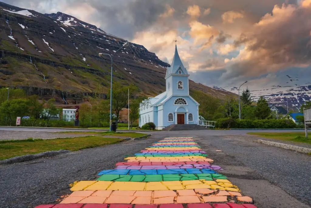 Colorful rainbow walkway leading to the iconic blue church in Seyðisfjörður, Iceland, surrounded by mountains and scenic landscape at sunset