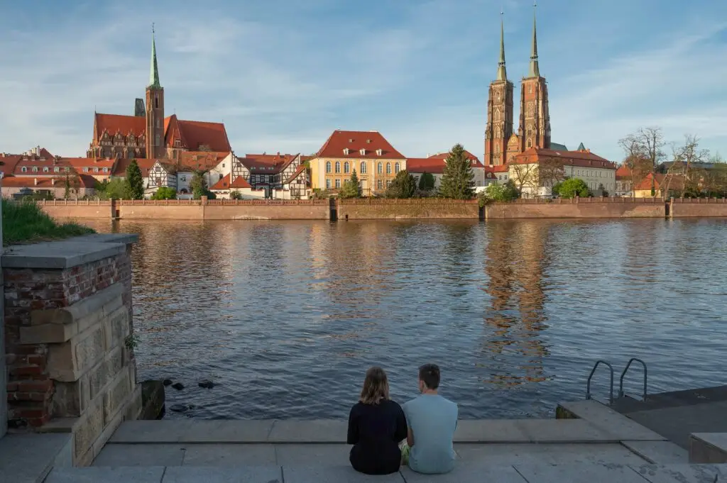 Couple sitting by the Oder River overlooking the historic cathedral and colorful buildings of Ostrów Tumski in Wrocław, Poland