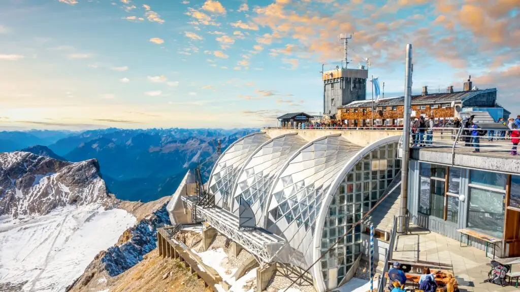 Panoramic view of Zugspitze mountain summit in Germany with visitors enjoying the observation deck, modern glass architecture, and alpine scenery in the Bavarian Alps.
