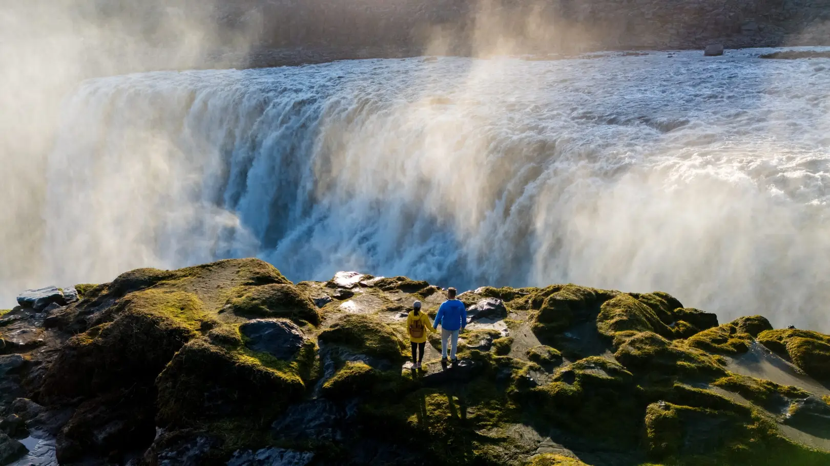 Travelers admiring the powerful Dettifoss Waterfall in North Iceland, surrounded by mist and rugged green cliffs, a popular destination for nature tours in Europe
