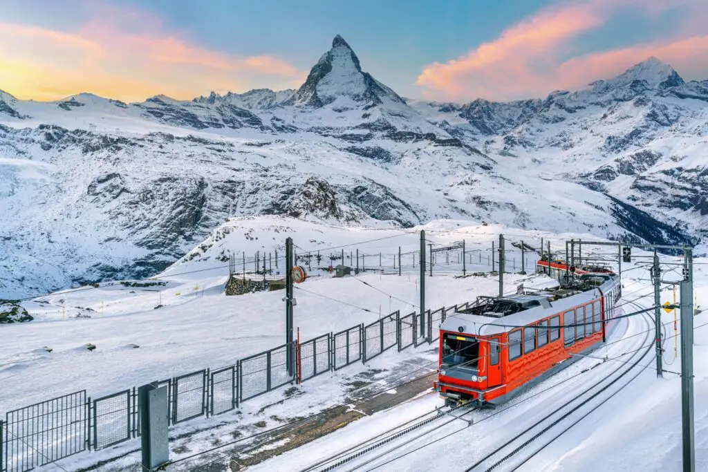 Red train traveling through snowy mountains near the Matterhorn in Zermatt, Switzerland at sunrise, showcasing scenic Swiss Alps winter landscape for Europe private tours