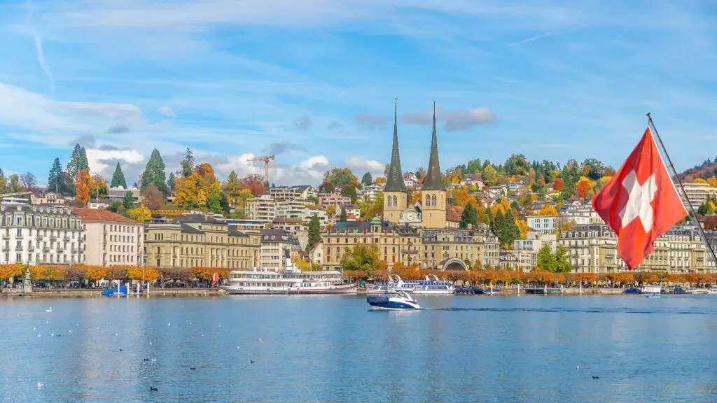 Scenic view of Lucerne, Switzerland with Lake Lucerne, historic architecture, church towers, boats, and Swiss flag on a bright autumn day