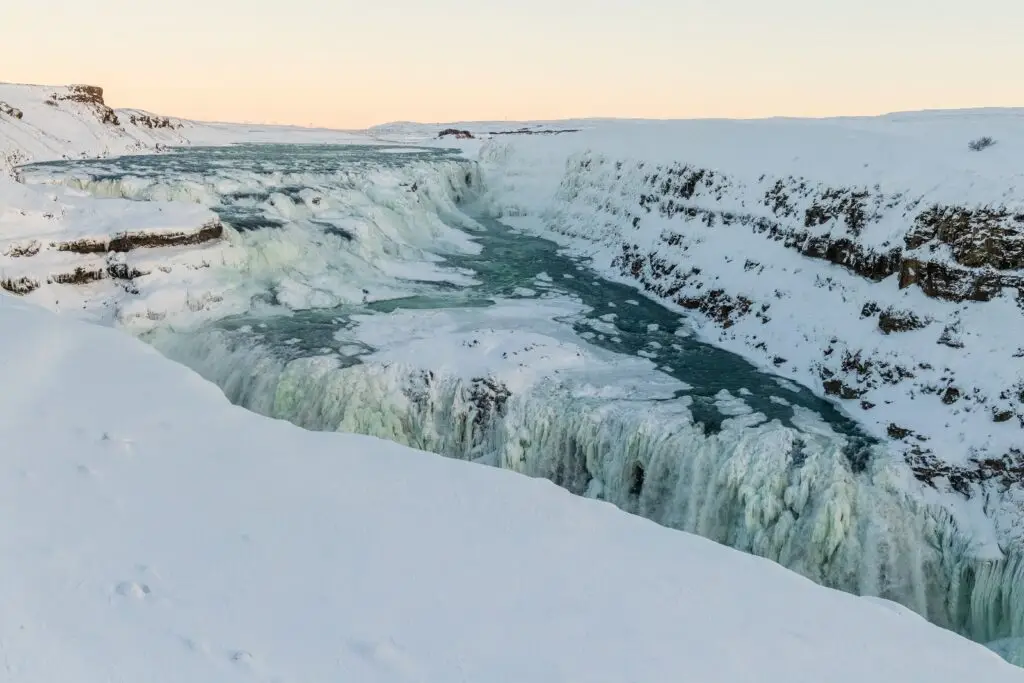 Gullfoss Waterfall in Iceland covered in winter snow and ice, showcasing powerful cascading waters and frozen cliffs at sunset, a popular stop on Iceland’s Golden Circle tours