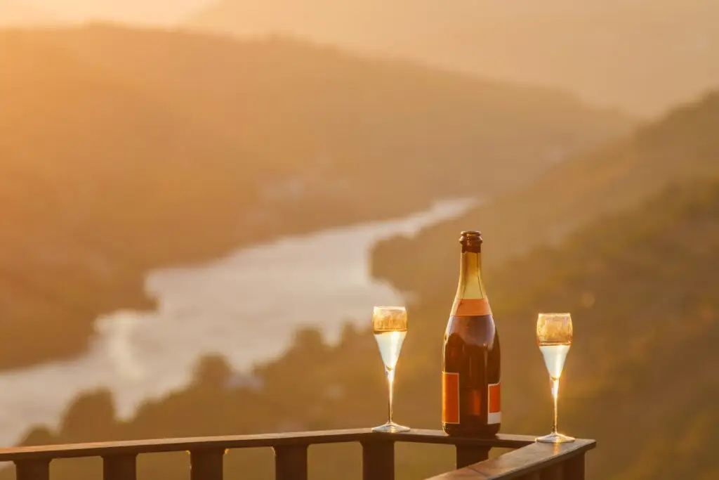 Bottle of sparkling wine and two glasses on a terrace overlooking a scenic European river valley at sunset