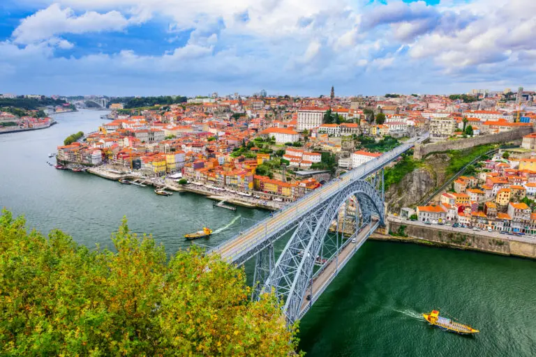 Panoramic view of Porto cityscape in Portugal featuring the Dom Luis I Bridge over the Douro River with colorful riverside buildings and boats.