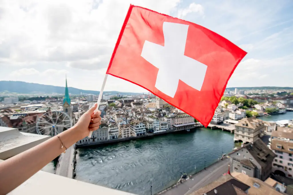 Person holding a Swiss flag overlooking the cityscape and river of Zurich, Switzerland on a sunny day, popular destination for European private tours