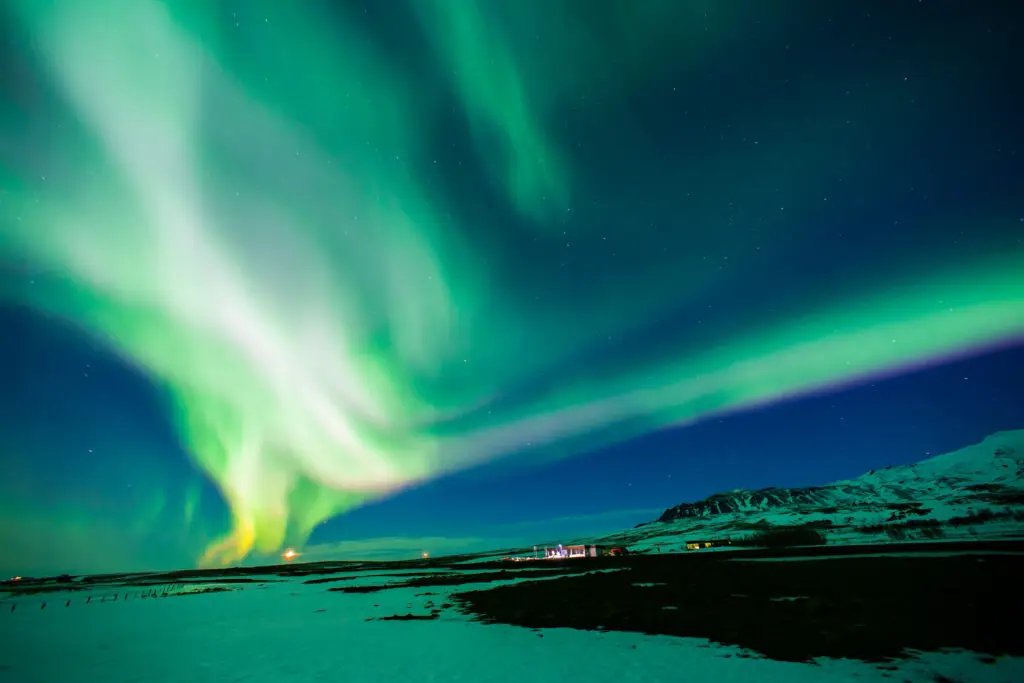 Northern Lights over snowy landscape in Iceland with mountains and rural houses illuminated under the night sky